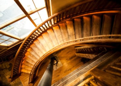 Close-up view of a curved wooden staircase winding upwards. The stairs are made of light-brown wood, appearing to be planks or wide boards.